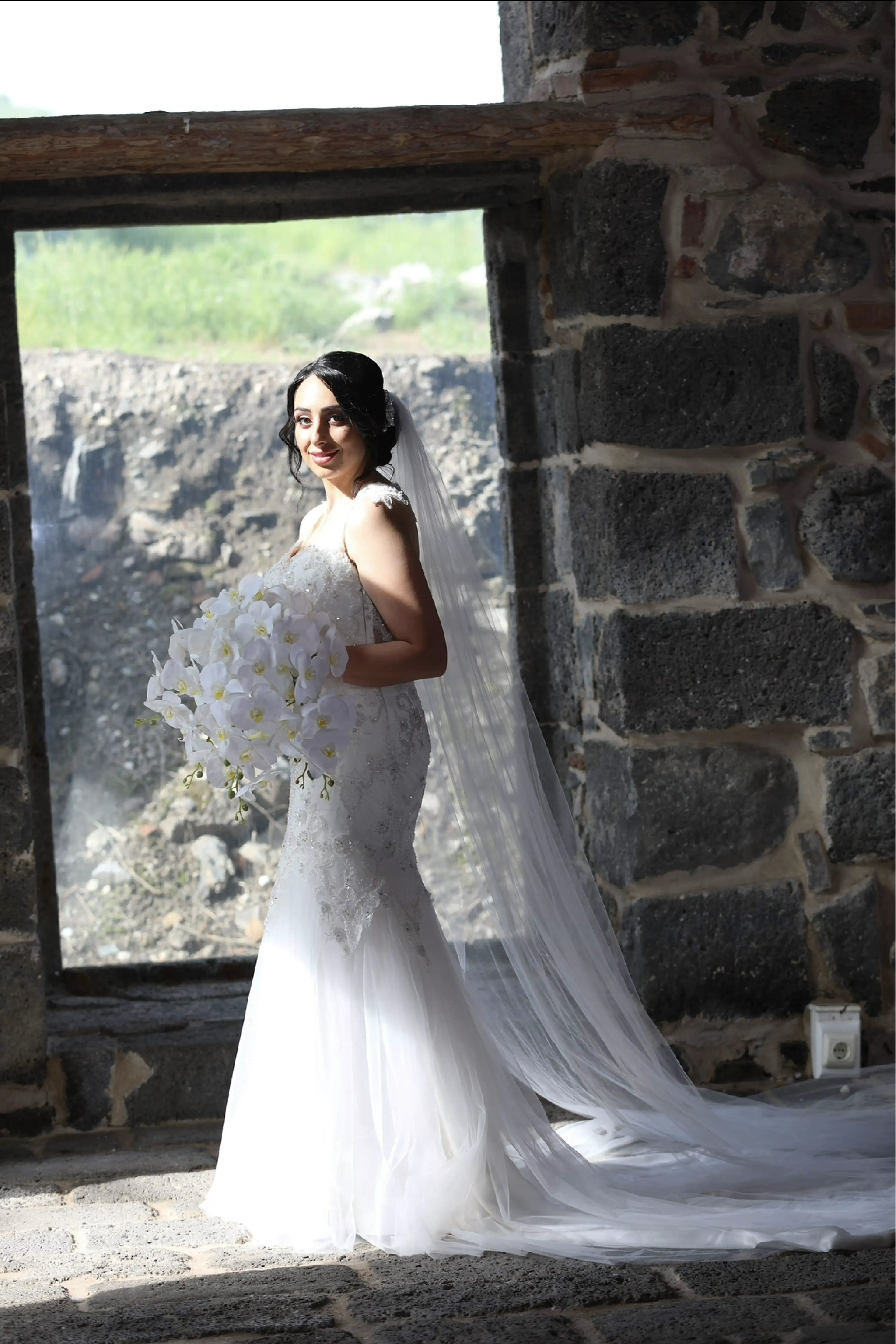 A bride in a lace wedding dress with a flowing headpiece, standing on a stone floor with her dress trailing behind her. She holds a bouquet of white flowers.