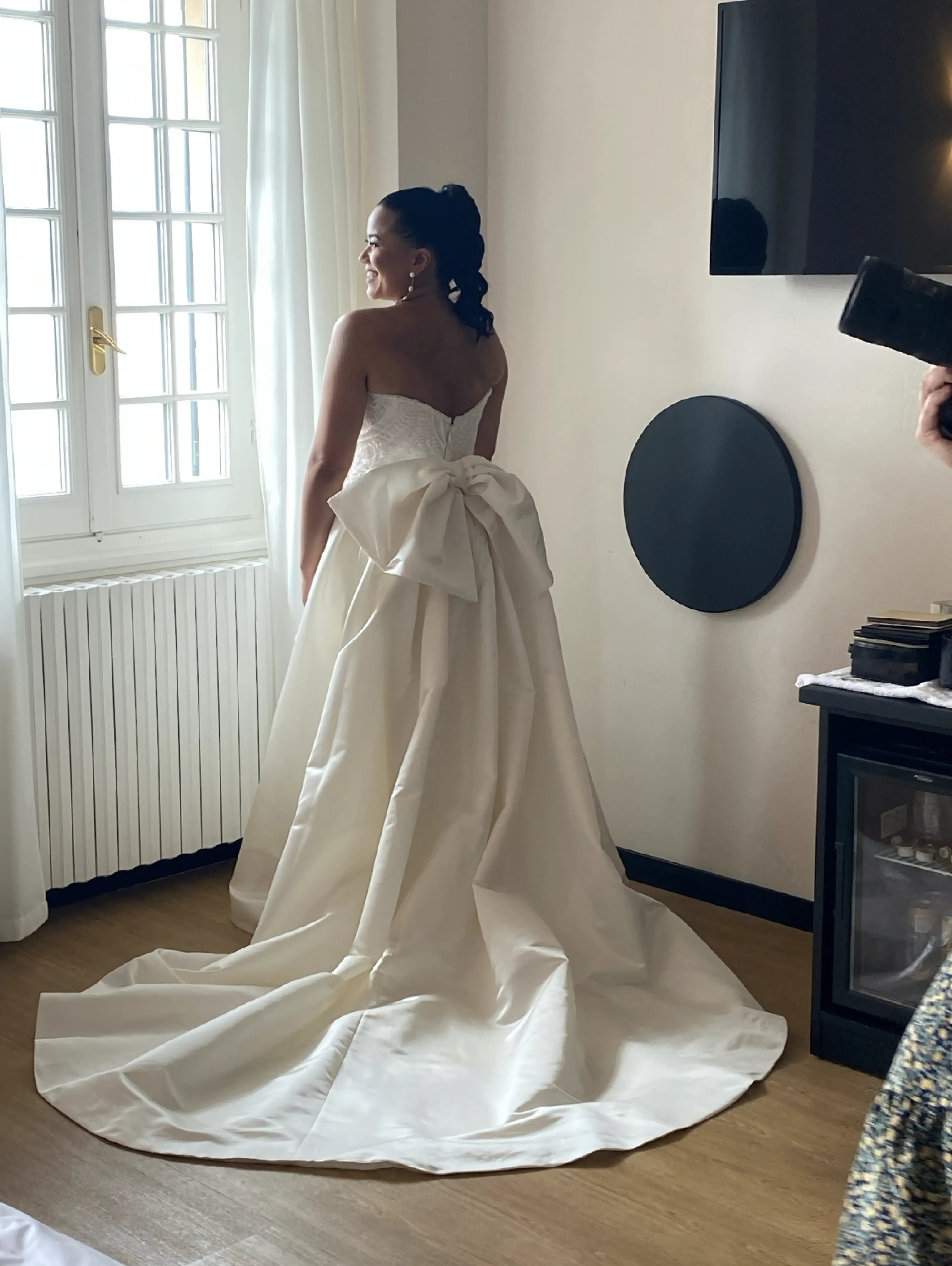 Bride with her back turned, wearing a wedding dress featuring a prominent silk bow at the center of her back. The fabric gracefully flows around the bow.